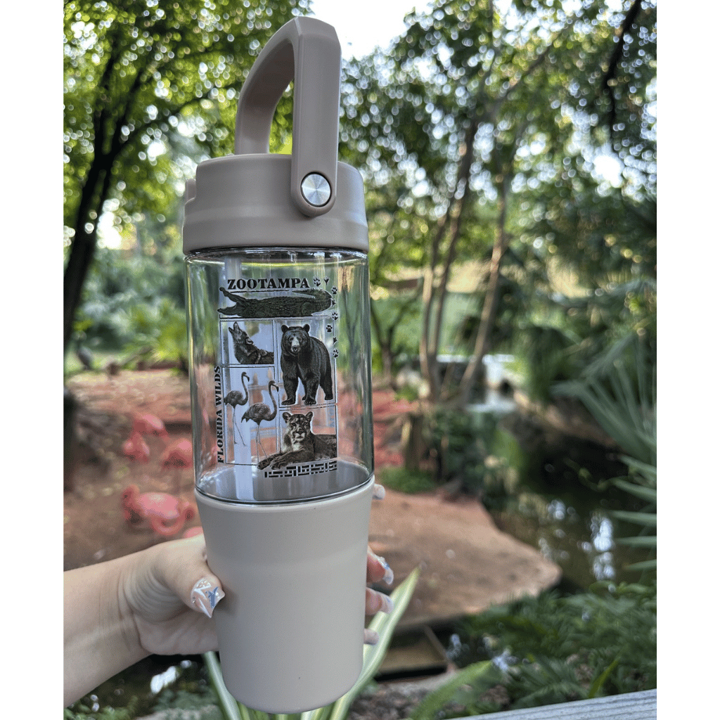 A clear plastic water bottle with a beige silicone lid and base, featuring a printed animal-themed label on the middle section. A hand is holding the bottle against a blurred outdoor background of trees and a pond.