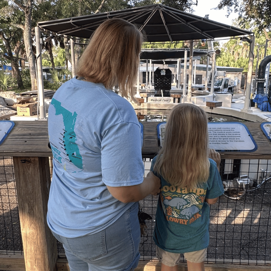 A woman and a young girl standing at a wooden exhibit railing, both wearing graphic t-shirts. The woman's light blue t-shirt features a teal map graphic of Florida, and the girl's teal t-shirt features a Florida manatee graphic with the text 'ZooTampa at Lowry Park'.