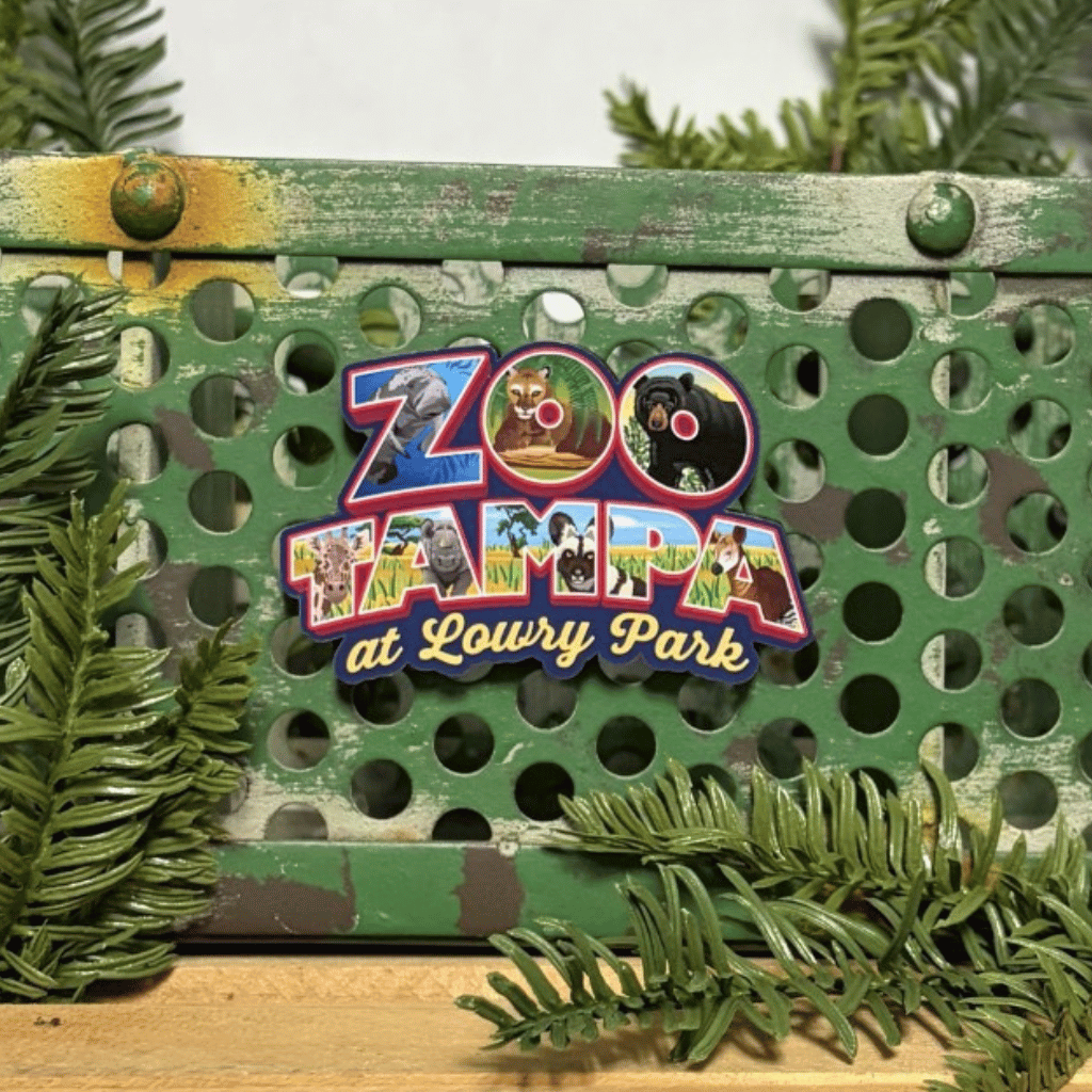 A colorful magnet shaped like a zoo map with the text 'ZooTampa at Lowry Park,' displayed on a distressed green metal crate with pine branches around it.