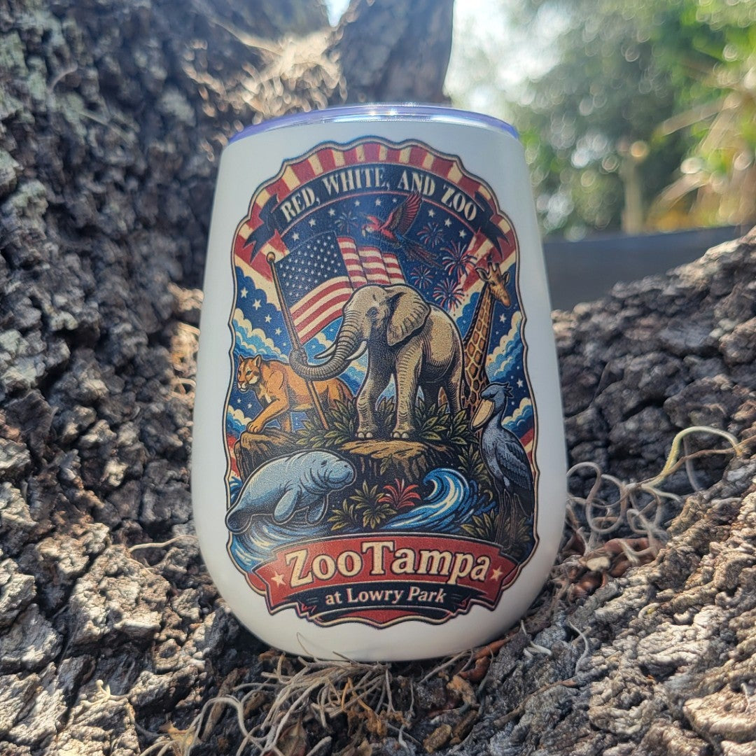 Red, White, and Zoo wine tumbler with animals holding a flag including elephant, giraffe, manatee, panther, shoebill. The cup is sitting on a tree stump.