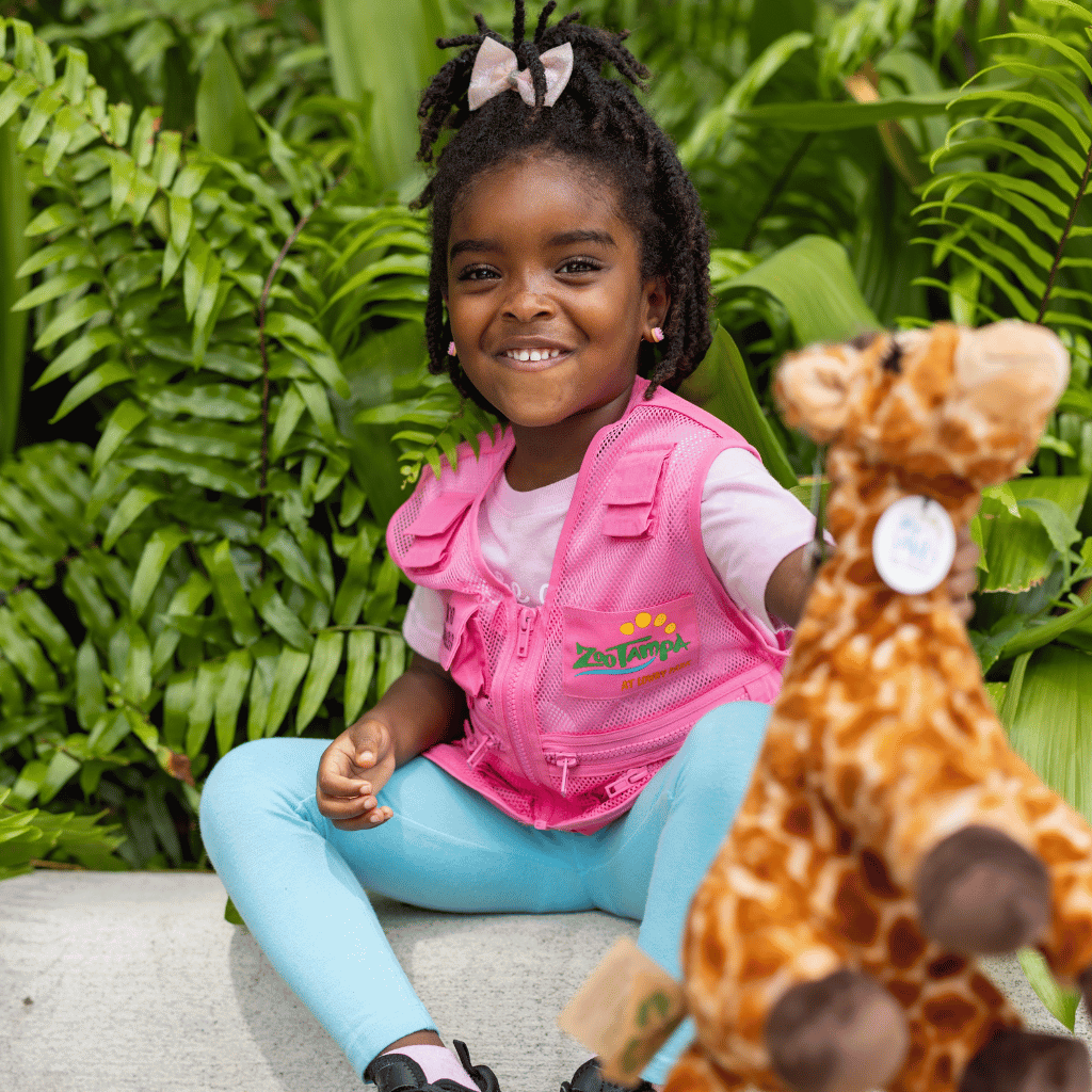 Little girl with twists wearing a pink zookeeper vest and holding a giraffe plush towards the camera