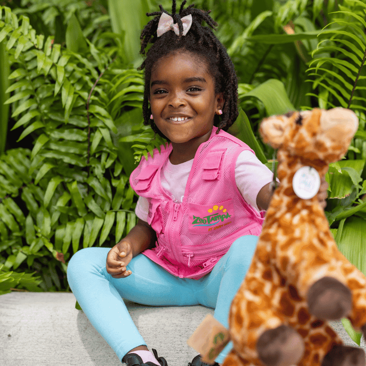 Little girl with twists wearing a pink zookeeper vest and holding a giraffe plush towards the camera