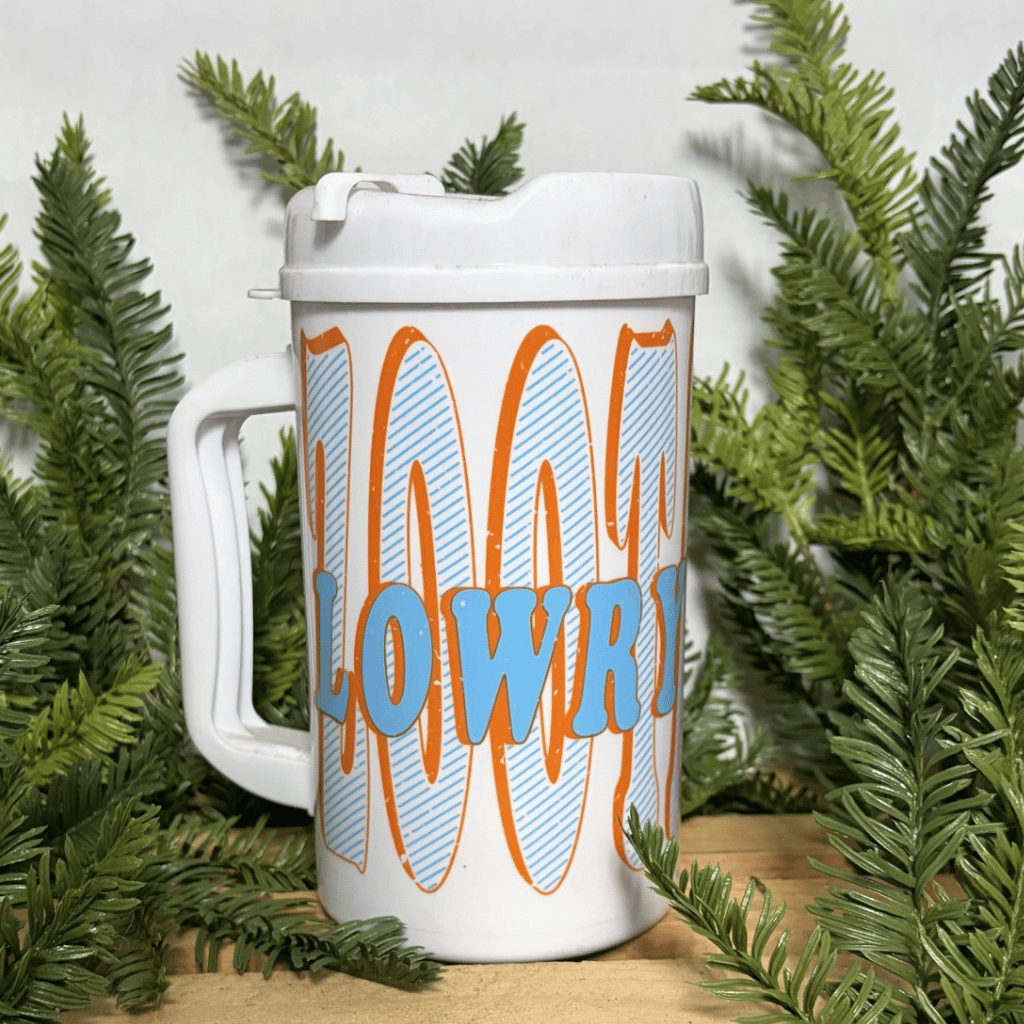 A white handled travel mug with a lid, featuring the words 'LOWRY PARK' in light blue letters over large orange and light blue striped words 'ZooTampa', styled with green branches on a wooden surface.