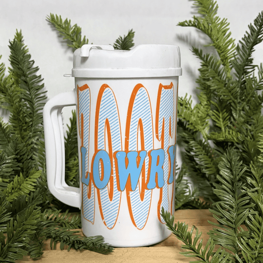 A white handled travel mug with a lid, featuring the words 'LOWRY PARK' in light blue letters over large orange and light blue striped words 'ZooTampa', styled with green branches on a wooden surface.