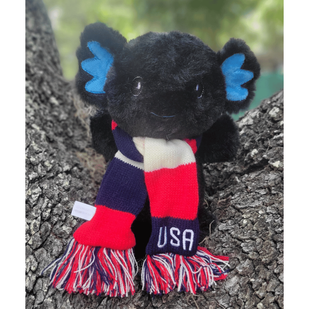 Black axolotl plush sitting on a tree stump wearing a red, white, and blue striped scarf with USA embroidered on it.