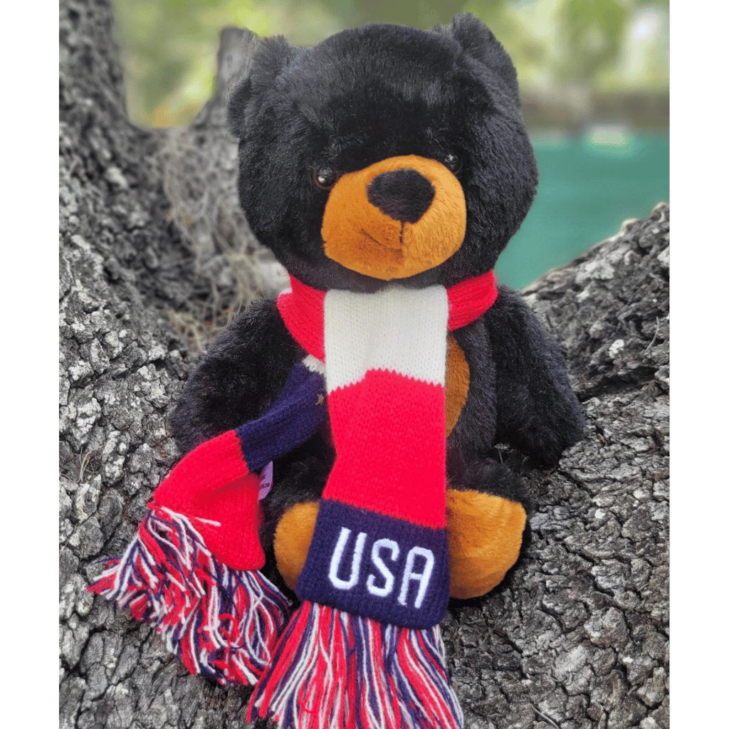 Black bear plush sitting on a tree stump wearing a red, white, and blue striped scarf with USA embroidered on it.