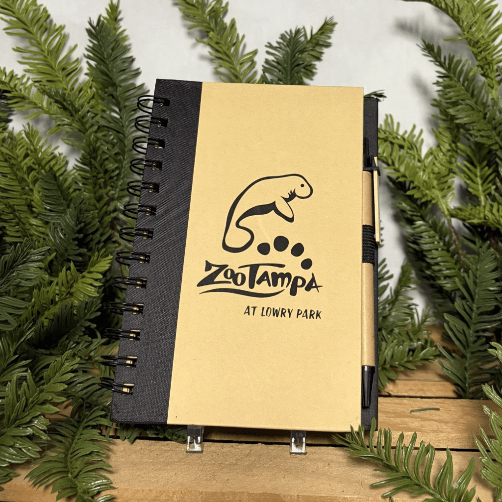 A spiral-bound notebook with a tan cover and black spine, featuring a manatee logo and the text 'ZooTampa At Lowry Park.' A black pen is tucked into the side, and the notebook is photographed among green pine branches on a wooden surface against a light background.