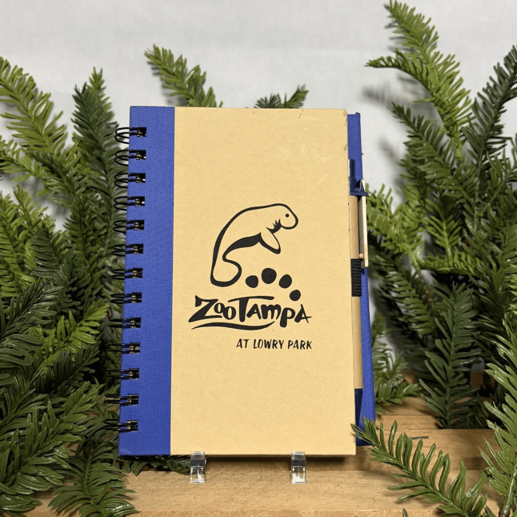 A spiral-bound notebook with a tan cover and blue spine, featuring a black manatee logo and the text 'ZooTampa At Lony Park.' It is displayed upright on small clear stands, surrounded by green pine branches on a wooden surface against a light gray background.