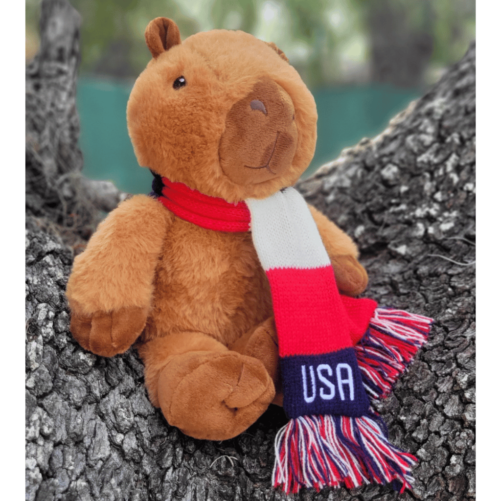 A brown plush capybara toy sitting on a tree trunk outdoors, wearing a red, white, and navy knit scarf with 'USA' embroidered on it.
