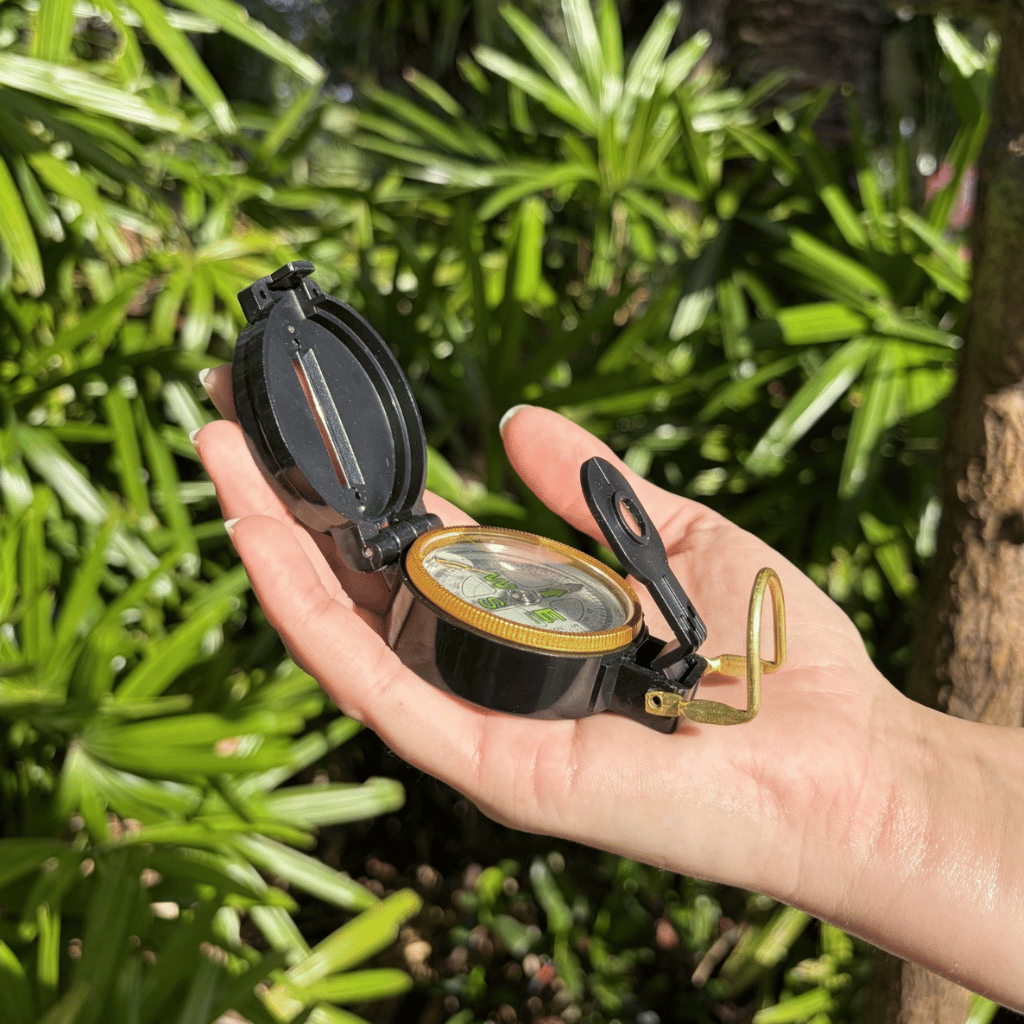 A black pocket compass with brass detailing is held in a person's hand, with a lush background of green palm leaves and a tree trunk.