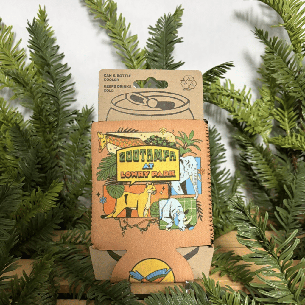 A beverage can cooler sleeve with colorful cartoon artwork reading 'ZooTampa at Lowry Park,' photographed upright among green pine branches against a light neutral background.