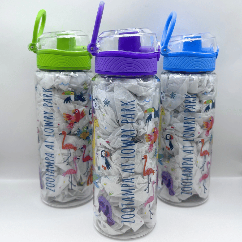 Three clear plastic water bottles with colorful lids and handles, filled with white paper crumpled into a textured pattern, printed with 'ZOO TAMPA AT LOWRY PARK' and tropical flamingo and toucan designs. The bottles have lime green, purple, and blue lids.