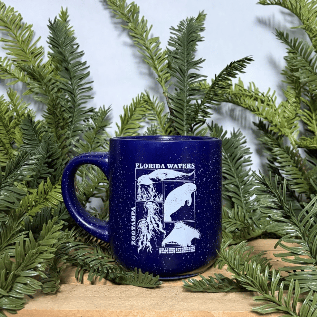 A dark blue speckled enamel mug with white graphics of marine life and the text 'FLORIDA WATERS' and 'ZooTampa', styled on a wooden surface surrounded by green pine branches against a white background.