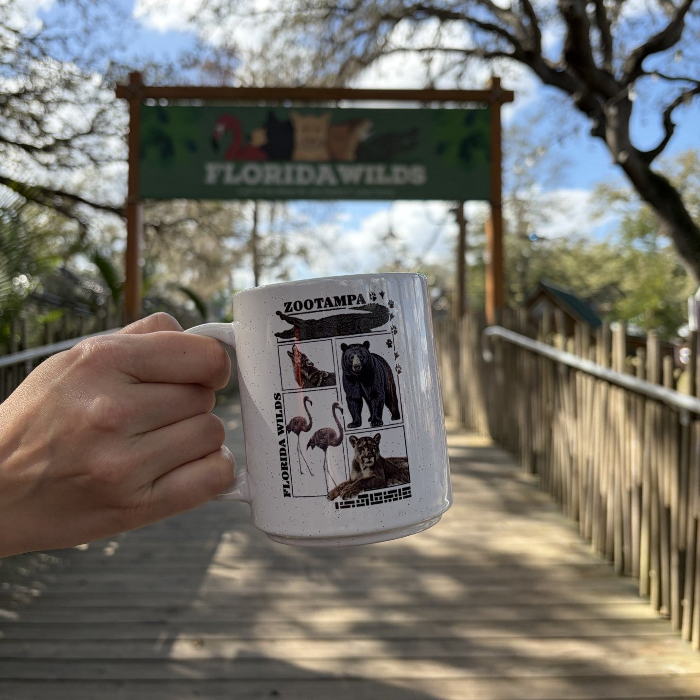 A hand holds a white ceramic mug printed with Florida Wildlife graphics, including a sign for Florida Wilds and animal photos, in front of a wooden boardwalk and Florida Wilds entrance under a blue sky with trees.