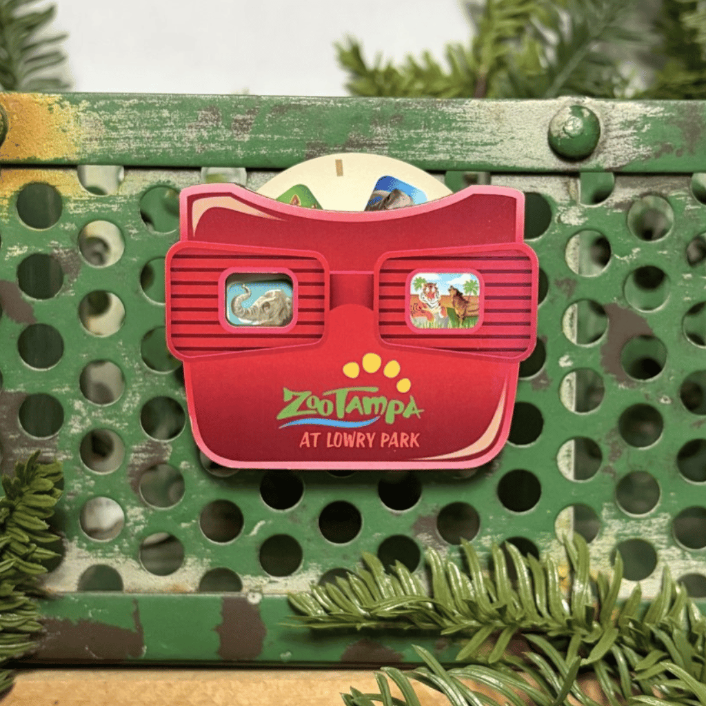 A red Zoo Tampa at Lowry Park 3D viewing glasses magnet with colorful cartoon scenes in the lenses, displayed on a distressed green metal surface with pine branches.