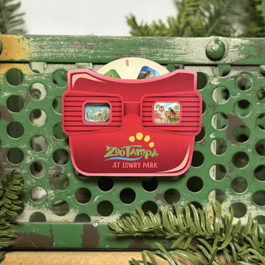 A red Zoo Tampa at Lowry Park 3D viewing glasses magnet with colorful cartoon scenes in the lenses, displayed on a distressed green metal surface with pine branches.