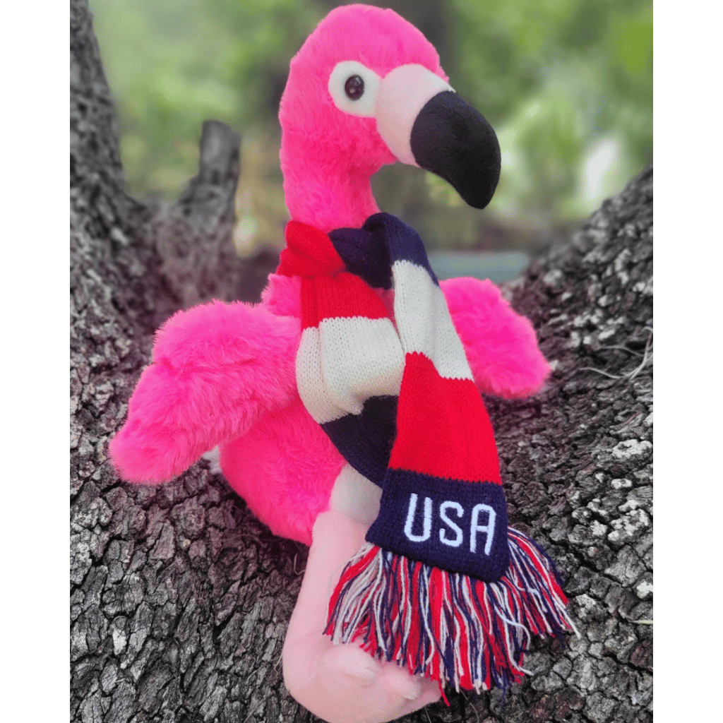 Flamingo plush sitting on a tree stump wearing a red, white, and blue striped scarf with USA embroidered on it.