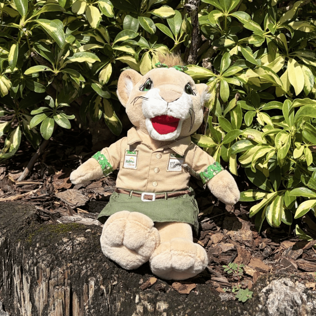 A plush ZooTampa panther mascot toy wearing a khaki safari-style shirt, green skirt, and green armband patches, sitting on a tree stump in front of green bush.