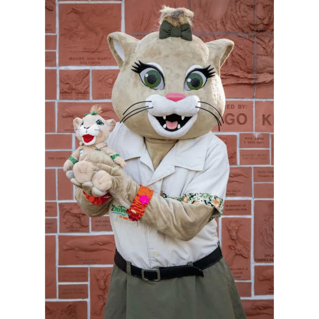 ZooTampa Flora the Panther Mascot stands in front of a red brick wall, holding a small plush toy. The costume includes a beige cat head with green eyes, a black bow, a white shirt, olive green skirt, and colorful wrist accessories.