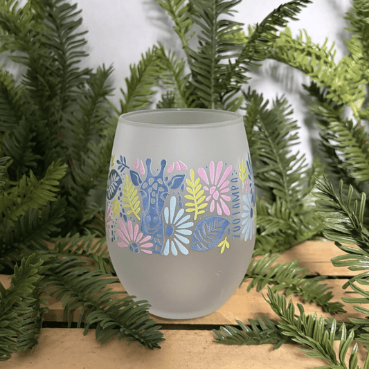 A frosted stemless wine glass with a colorful giraffe and floral pattern, displayed on a wooden surface surrounded by green pine branches.