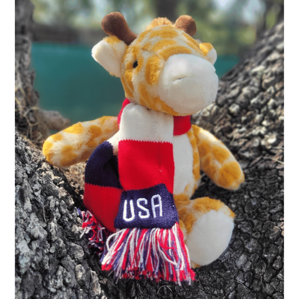 A plush giraffe toy sitting on tree bark outdoors, wearing a red, white, and navy USA-themed scarf with fringe and a small 'USA' patch.