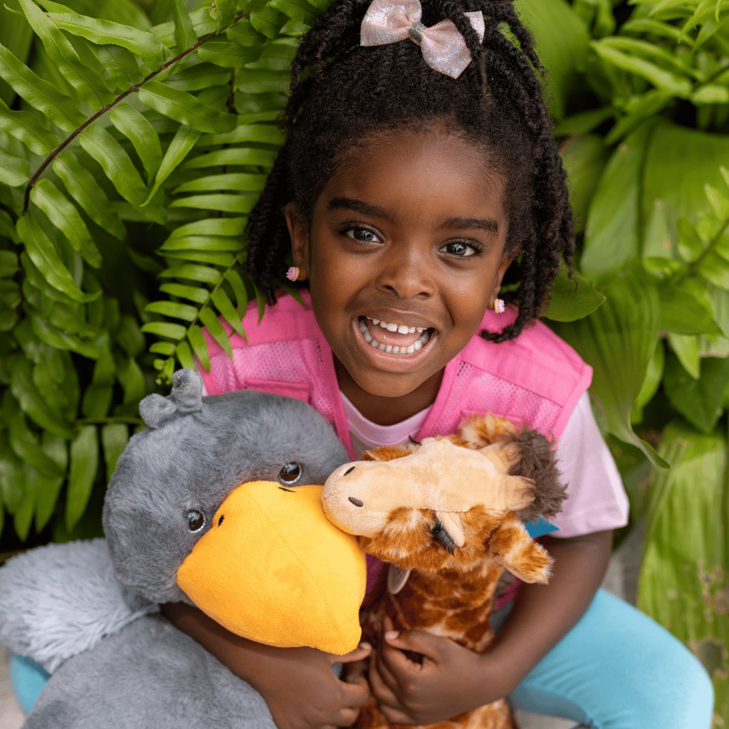A young girl with braided hair and a pink bow smiles while holding two plush toys, a gray bird with a yellow beak and a giraffe, in front of a background of green plants.