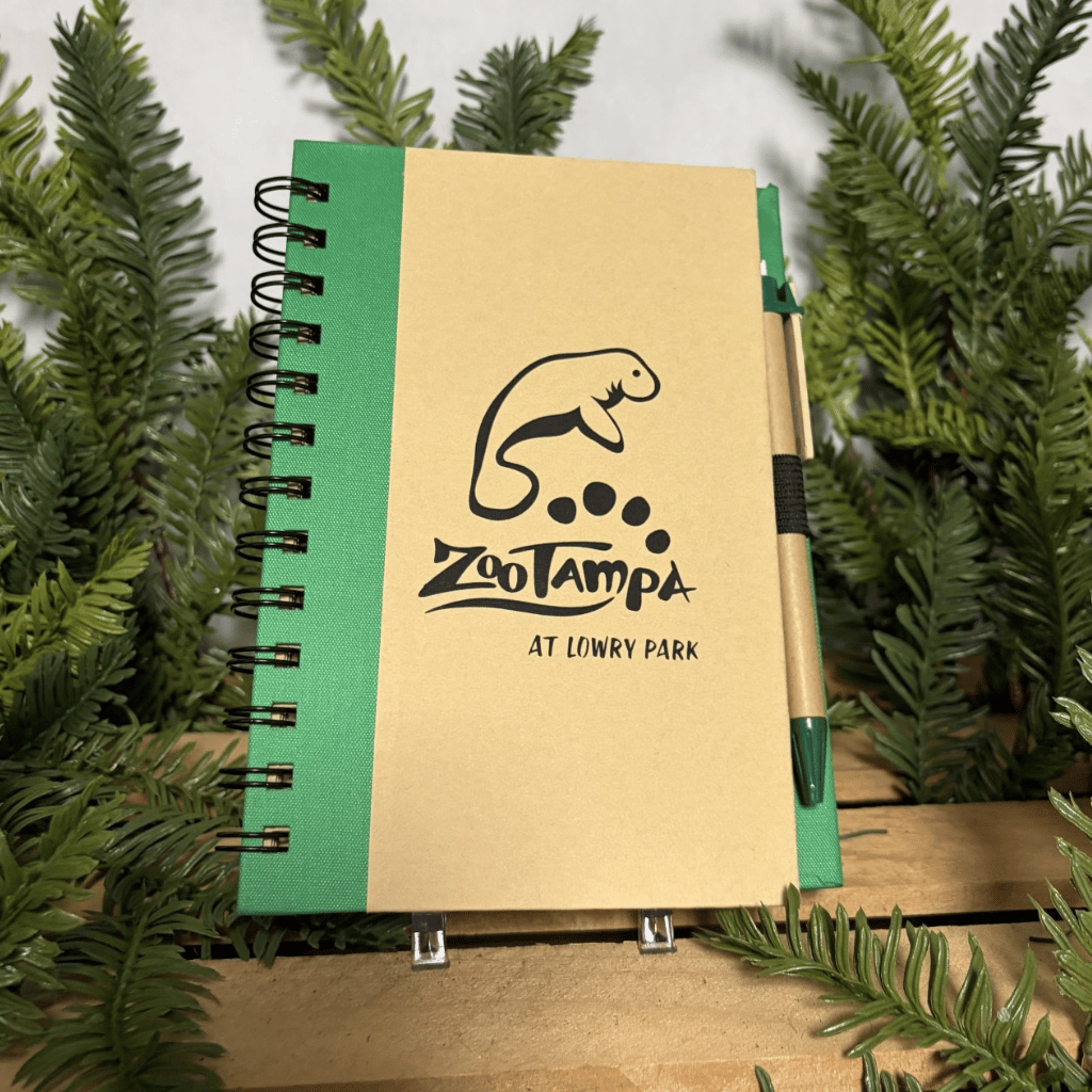A spiral-bound notebook with a tan cover and green spine, featuring a black zebra illustration and the text 'ZooTampa at Lowry Park.' A matching pen is tucked into the side, and the notebook is photographed among green pine branches on a wooden surface.