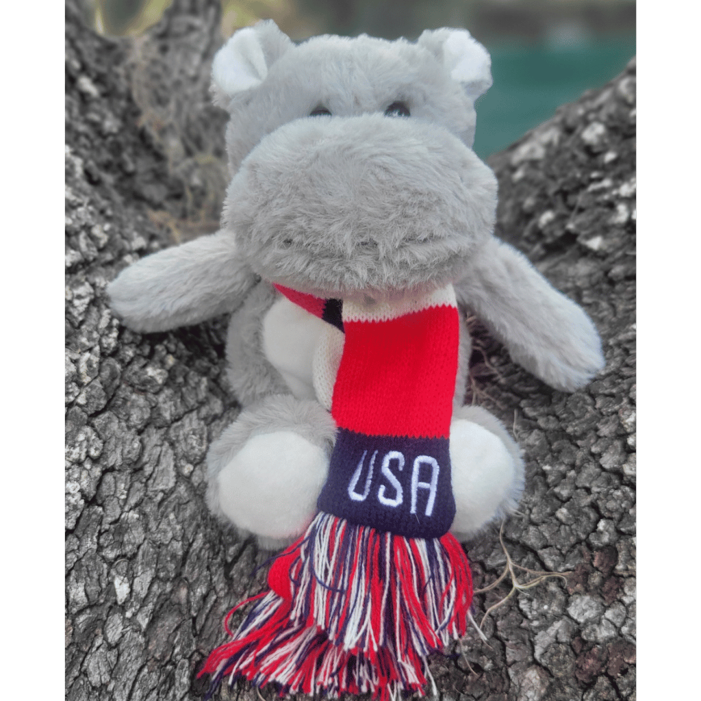 A Hippo plush sitting on a tree stump wearing a red, white, and blue striped scarf with USA embroidered on it.