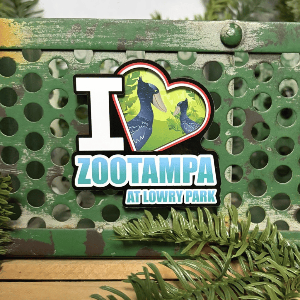 A die-cut magnet featuring the text ' ZooTampa at Lowry Park' with a heart-shaped illustration of shoebill storks, displayed on a green metal bench with pine branches in the foreground.
