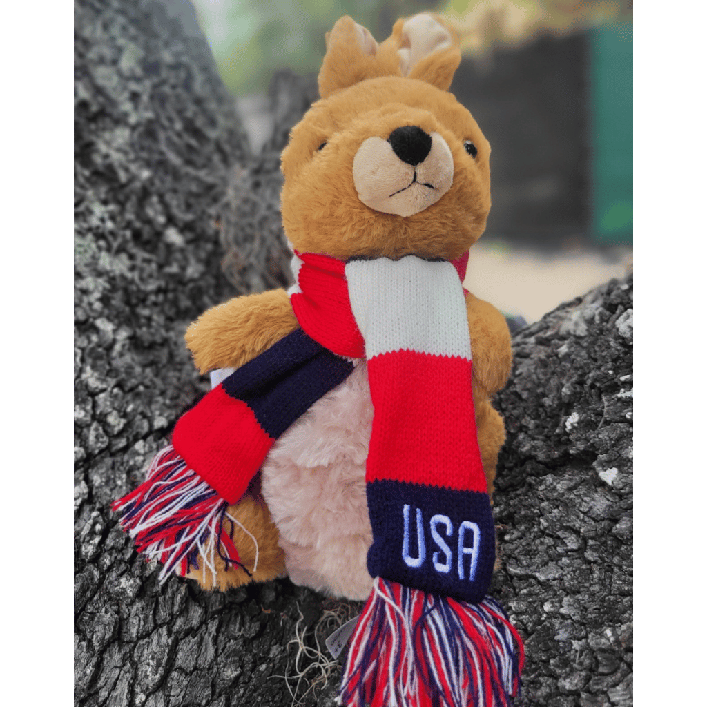 A plush kangaroo or wallaby wearing a red, white, and navy striped scarf with 'USA' embroidered on the navy end, posed against a tree trunk outdoors.