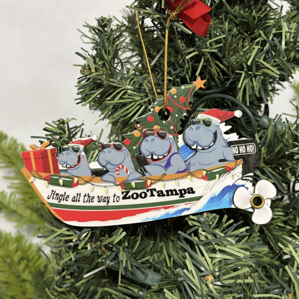 A Christmas ornament shaped like a boat carrying four cartoon manatees wearing Santa hats, hanging on a green artificial Christmas tree. The ornament reads 'Jingle all the way to ZooTampa' and has a red hanging ribbon.