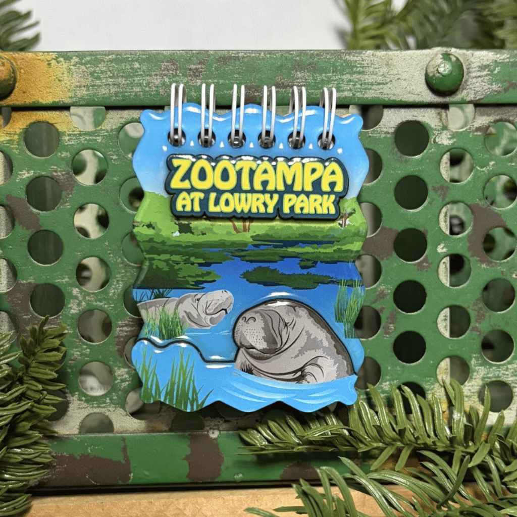 A ZooTampa at Lowry Park mini notebook magnet featuring an illustration of a manatee in a pond, displayed on a green perforated metal surface with pine branches.