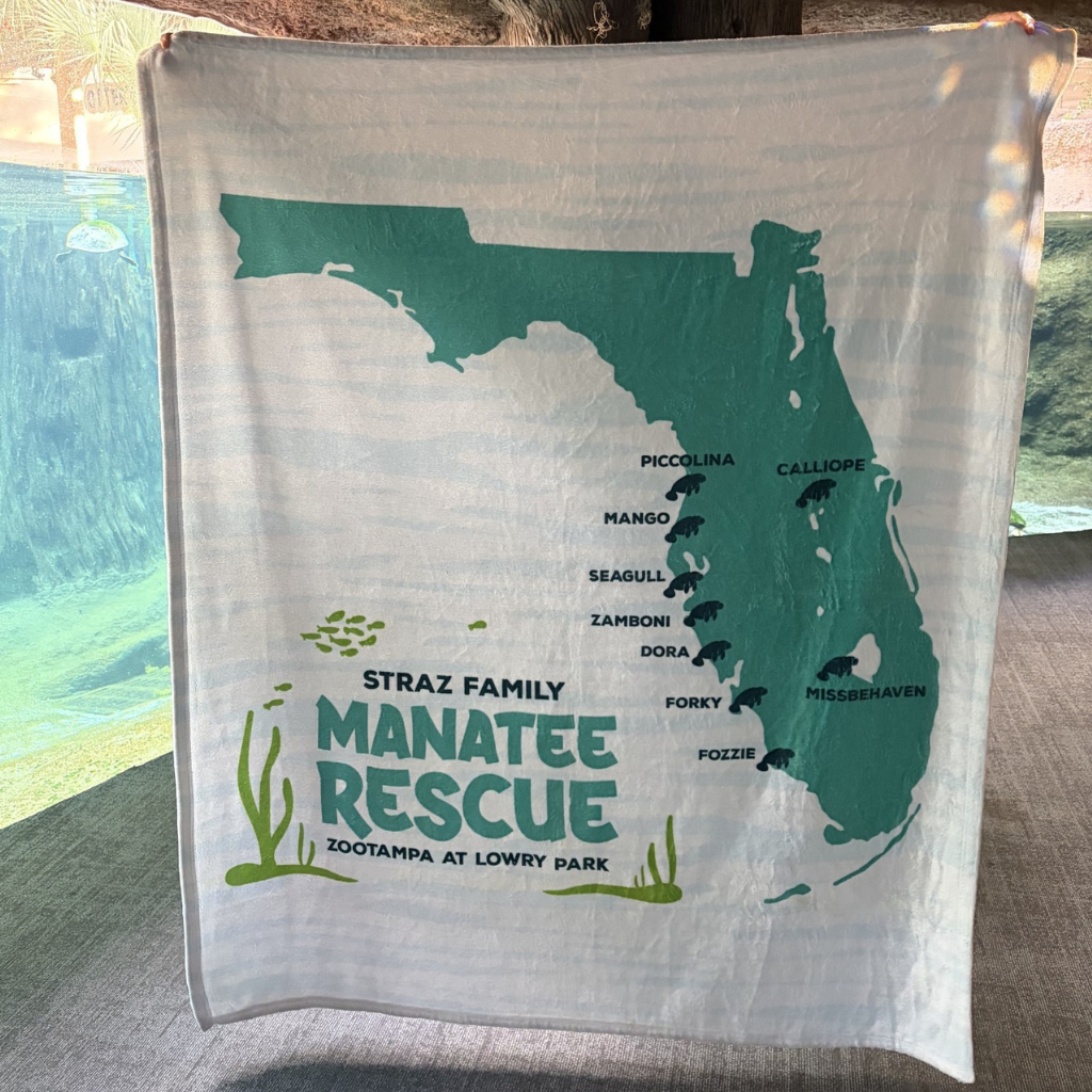 A white blanket featuring a teal map of Florida with the text 'Straz Family Manatee Rescue' and 'ZooTampa at Lowry Park,' displayed outdoors near a pool.