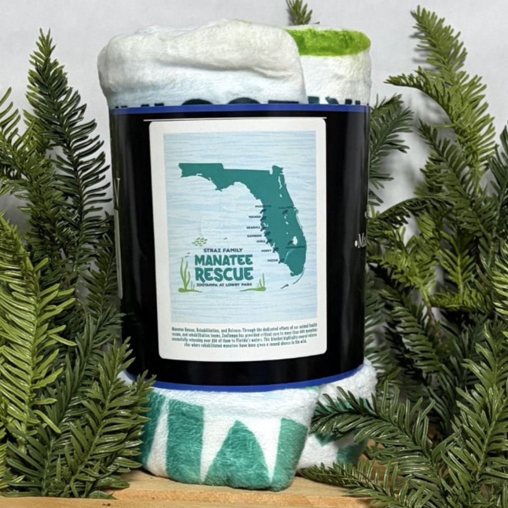 A rolled white blanket with green and white patterned ends, featuring a Florida map and the words 'Manatee Rescue.' Green pine branches surround the blanket against a light gray background.