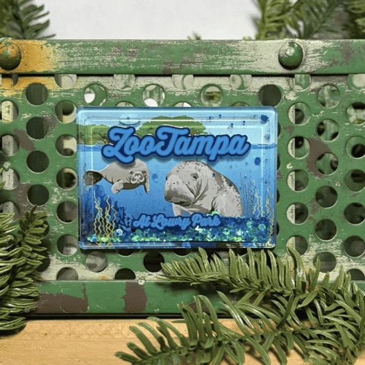A rectangular blue magnet featuring 'ZooTampa at Lowry Park' with illustrations of manatees, floating sparkles, displayed on a distressed green metal surface with pine branches around it.