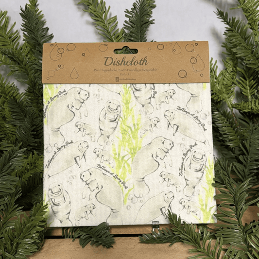 A packaged dishcloth with a kraft paper header card and a white cloth printed with black line drawings of manatee and light green watercolor accents, displayed among green pine branches.