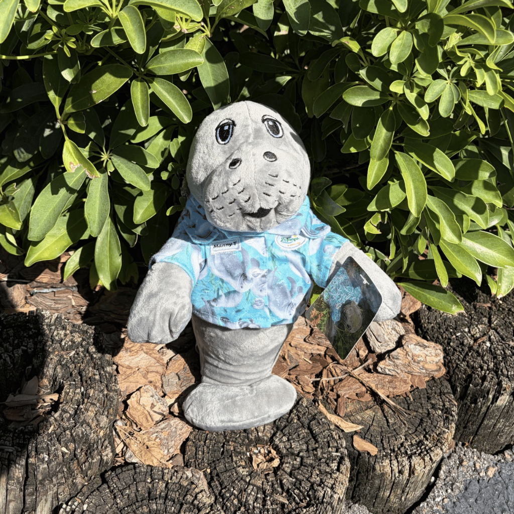 ZooTampa's manatee mascot - Manny T in plush form, wearing a blue and white tie-dye shirt, standing on a tree stump in front of green bushes