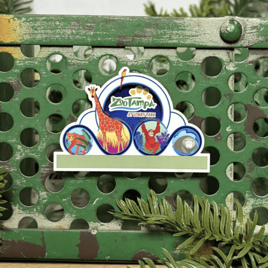 A ZooTampa magnet shaped like the zoo's front entrance marque, displayed on a distressed green metal shelf with pine branches in the foreground.