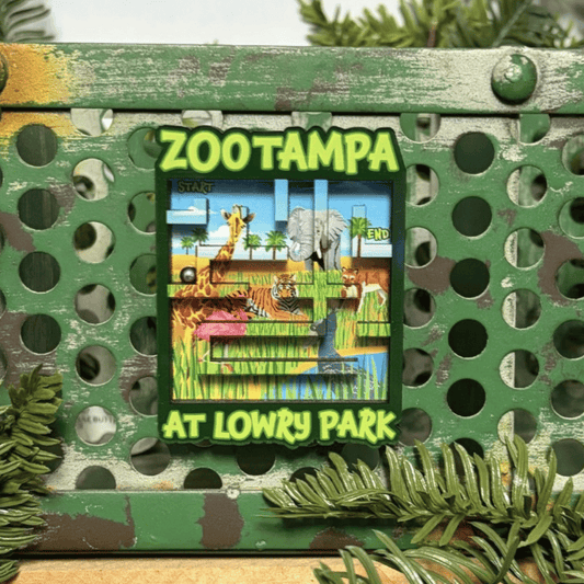 A square Zoo-themed magnet reading 'ZooTampa at Lowry Park' with a colorful safari scene in the center, mounted on a distressed green metal frame with decorative pine branches around it.