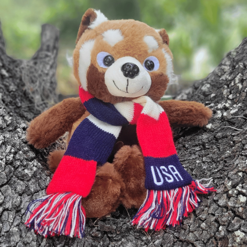 Red panda plush sitting on a tree stump wearing a red, white, and blue striped scarf with USA embroidered on it.