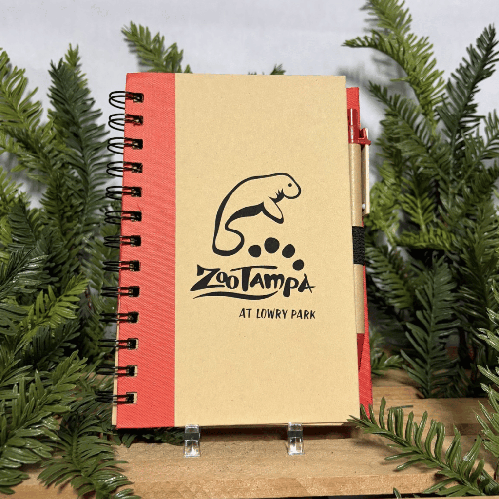 A spiral-bound notebook with a kraft paper cover, coral red spine, and black spiral binding, standing on clear acrylic blocks. The cover features a black manatee logo and the text 'ZooTampa at Lowry Park'.