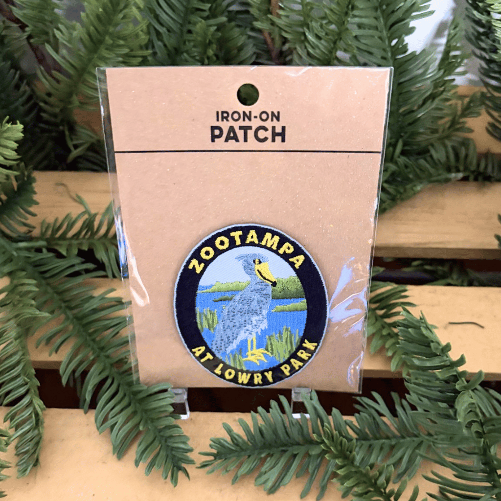 A round iron-on patch with a shoebill stork and words 'ZooTampa at Lowry Park' displayed on its kraft paper backing card, surrounded by green pine branches on a wooden surface.