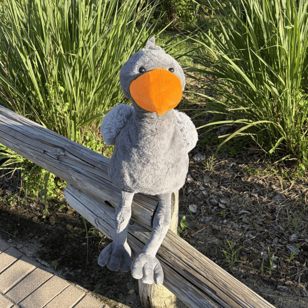A plush gray shoebill toy with a bright orange beak sits on a weathered wooden fence outdoors, with tall green grass and mulch in the background.