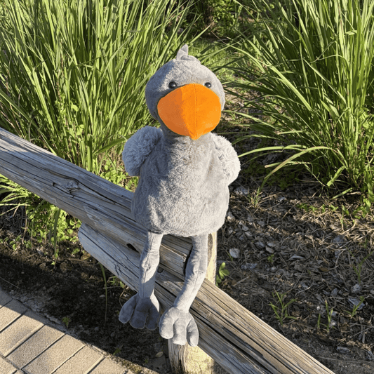 A plush gray shoebill toy with a bright orange beak sits on a weathered wooden fence outdoors, with tall green grass and mulch in the background.