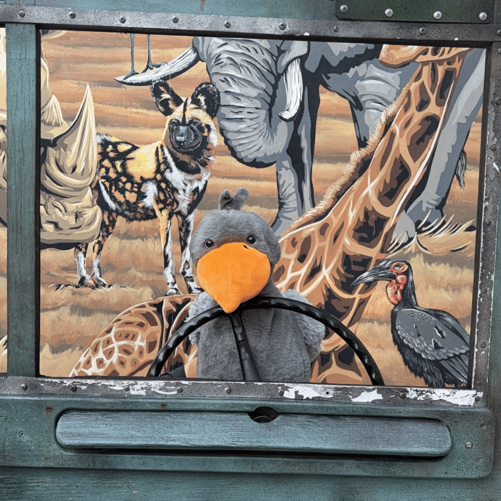 A plush toy shoebill with a bright orange beak and gray body, positioned in front of a painted safari-themed backdrop featuring an elephant, giraffe, rhino, and bird.