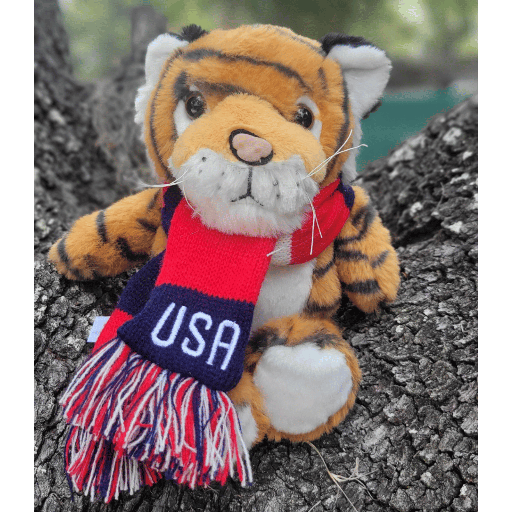 A plush tiger toy sitting on a tree trunk outdoors, wearing a red, navy, and white USA scarf with fringe.