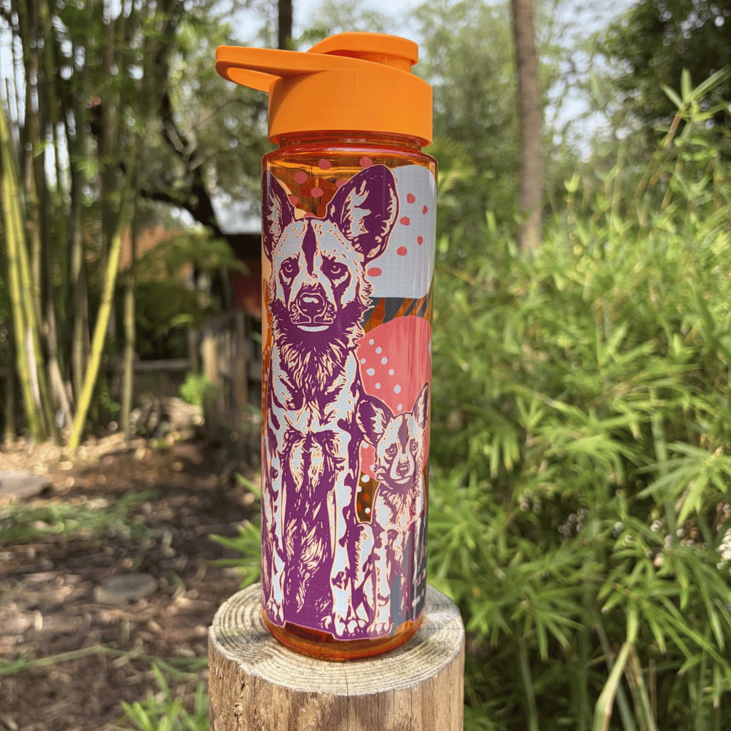 An orange flip-top water bottle with a colorful wild dog illustration on the front, standing on a wooden stump in an outdoor garden setting with bamboo in the background.