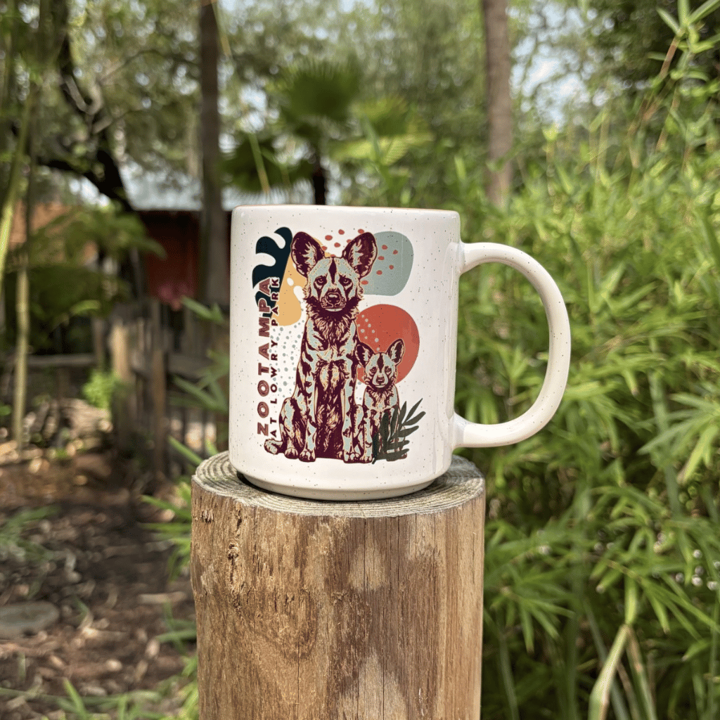 A white ceramic mug featuring a colorful illustration of two African Painted Dogs and the text ZooTampa at Lowry Park with blurred green foliage in the background.