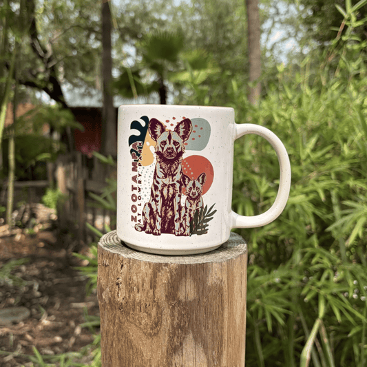 A white ceramic mug featuring a colorful illustration of two African Painted Dogs and the text ZooTampa at Lowry Park with blurred green foliage in the background.