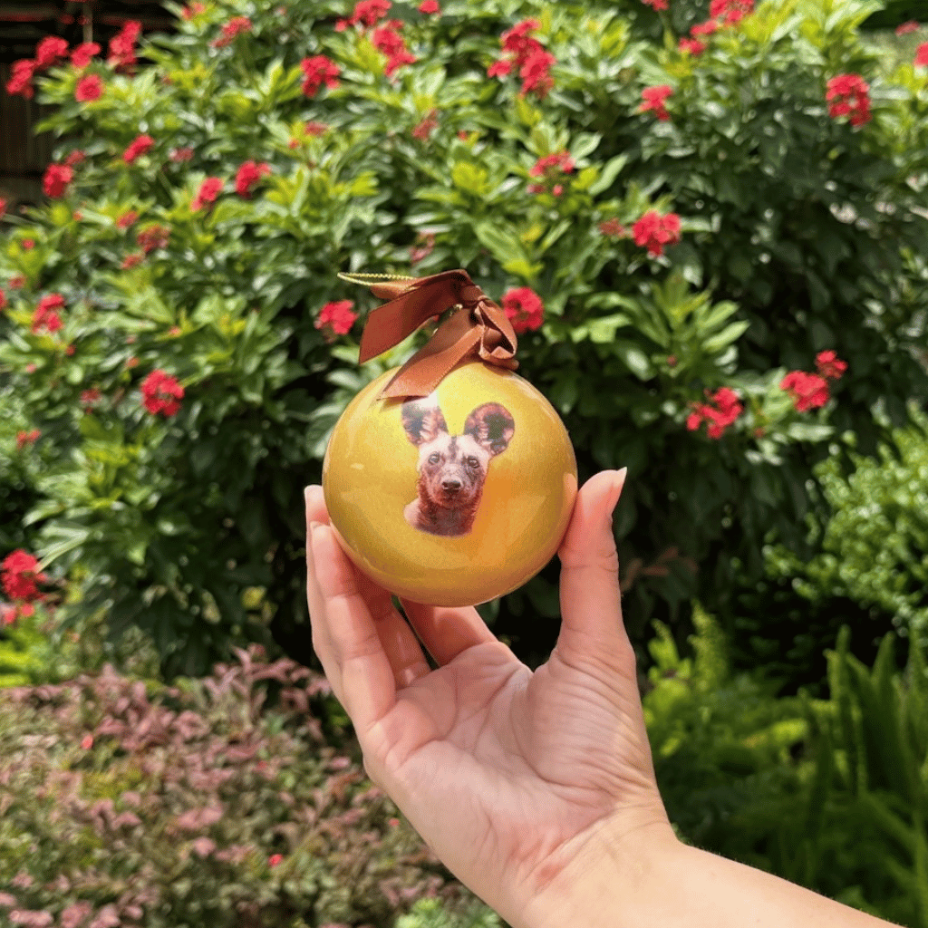 A hand holds a round gold Christmas ornament featuring a painted portrait of a dog, with a brown ribbon loop at the top, set against a background of green foliage and red flowers.